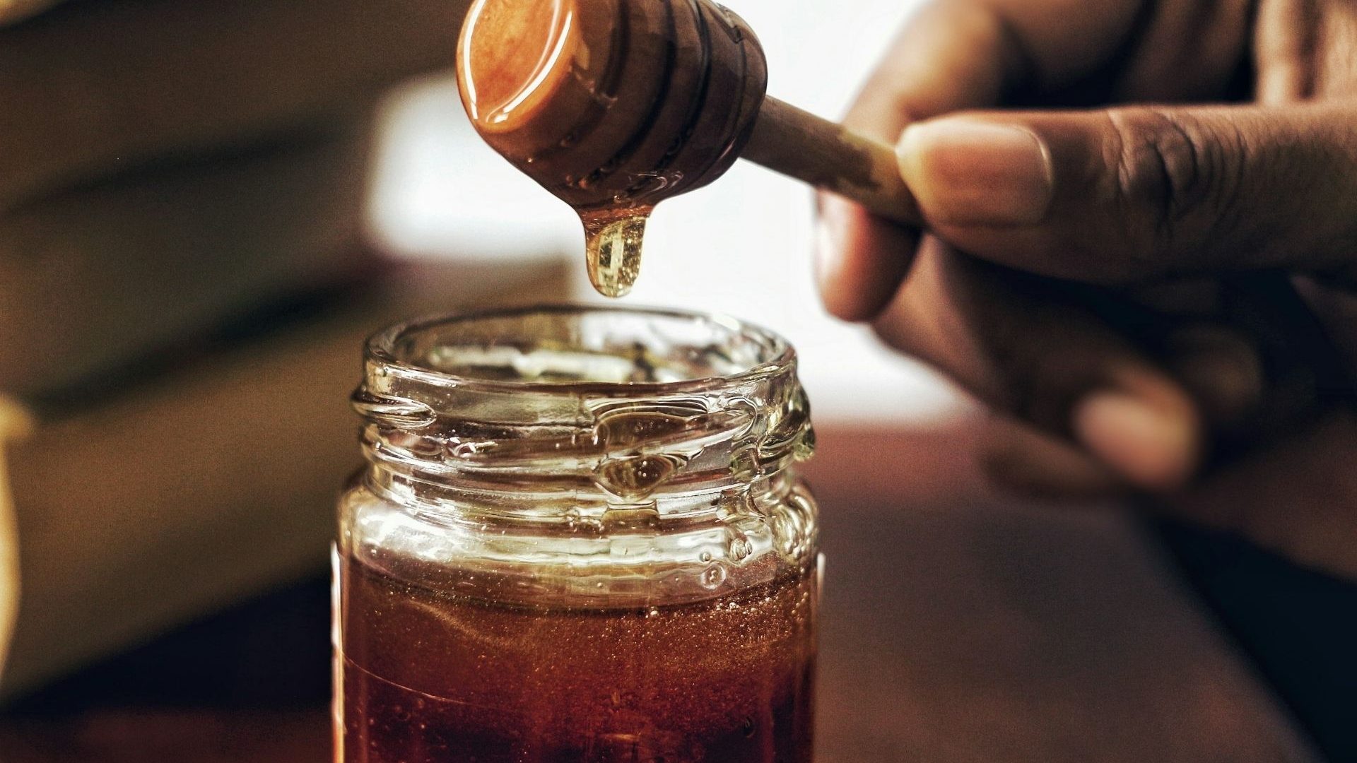 A hand holding a wooden honey dipper above a jar full of honey