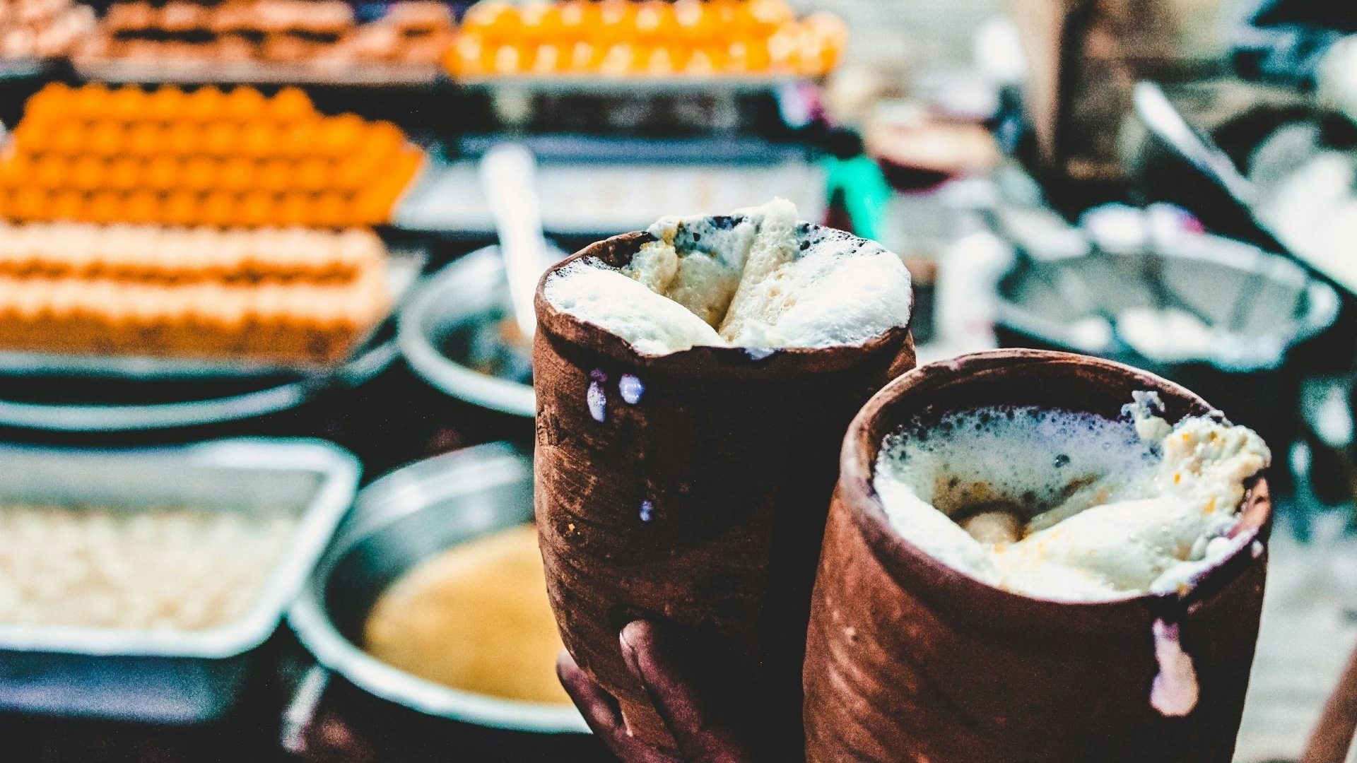 Two traditional wooden mugs of lassi yogurt drink against a blurred-out backdrop of Indian sweets and snacks