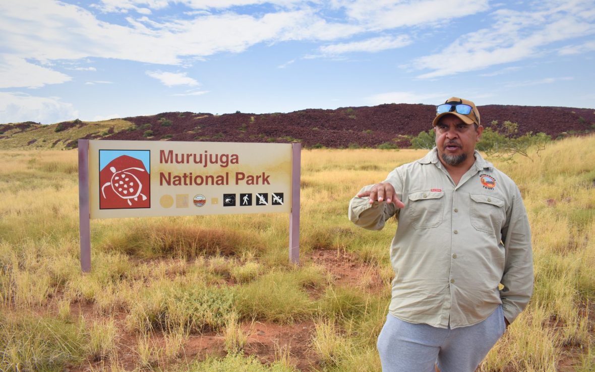 Male guide stands in an open field in front of a sign reading 'Murujuga National Park'