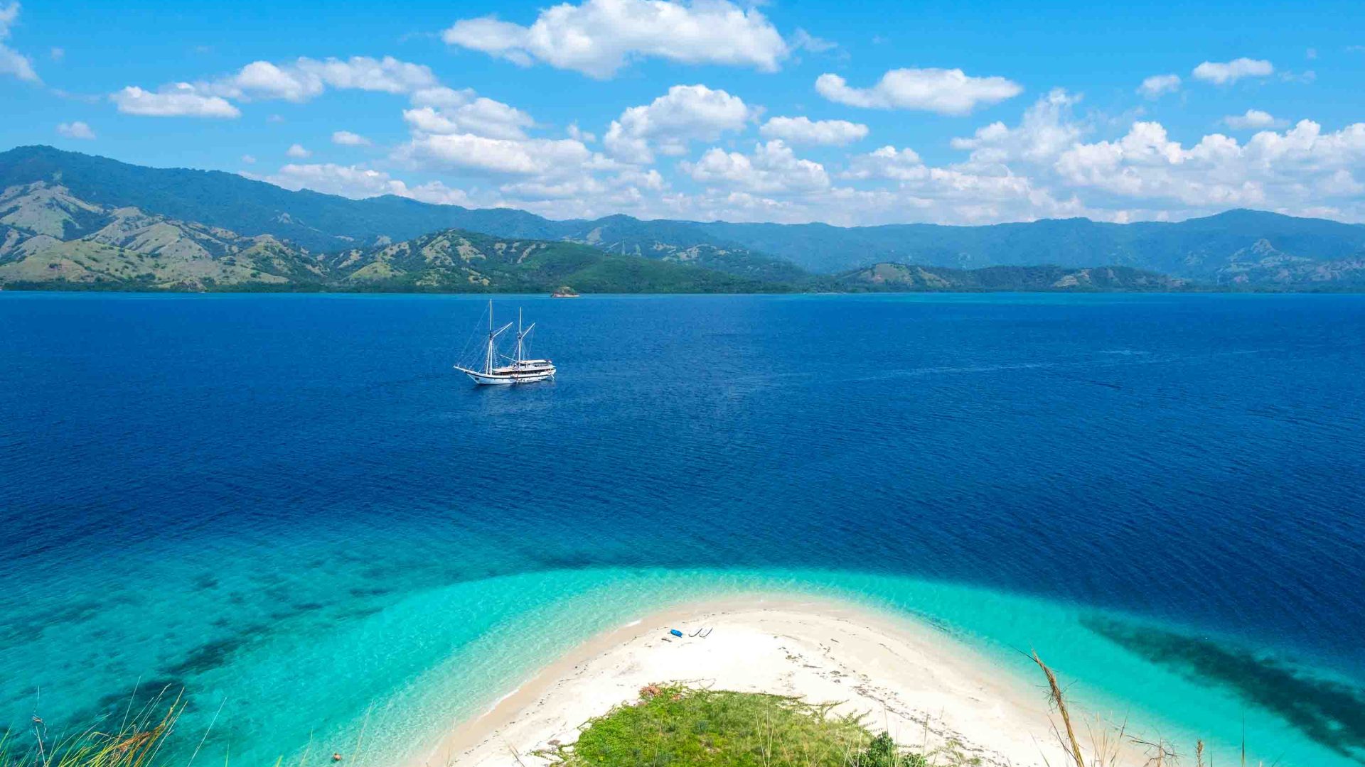 A boat in blue water with a beach in the foreground with white sand and turquoise water.