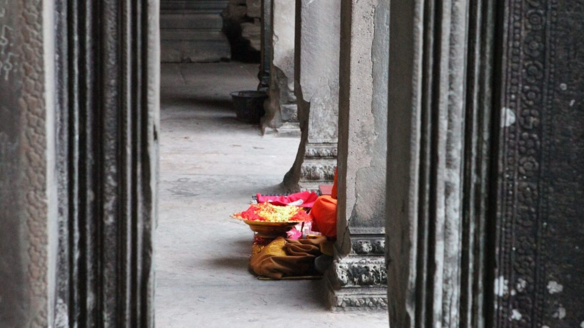 Against the pillars inside a temple complex, a glimpse of someone sat down with prayer offering of flower petals in their lap.