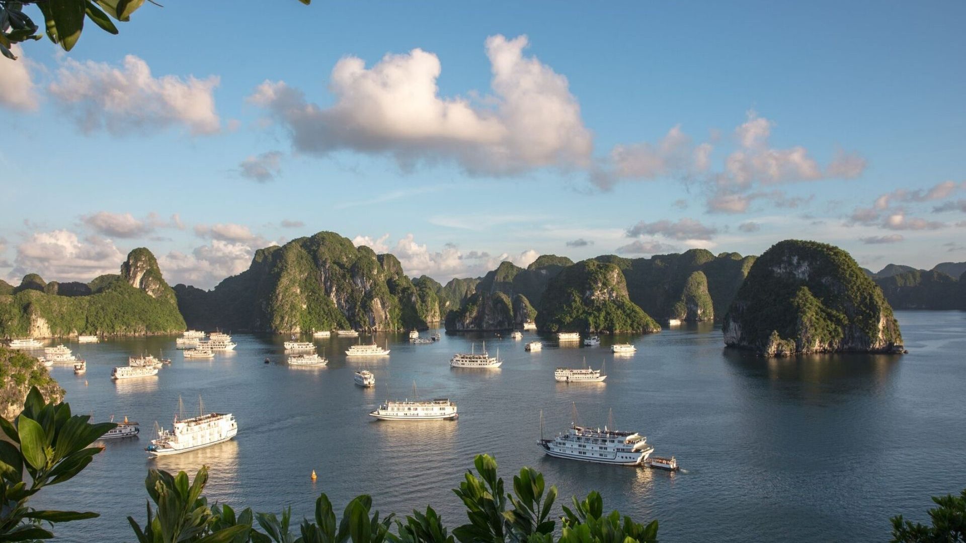 A bucolic scene of boats on the water at Ha Long Bay, against limestone karsts and forest, and a blue sky with a handful of clouds.