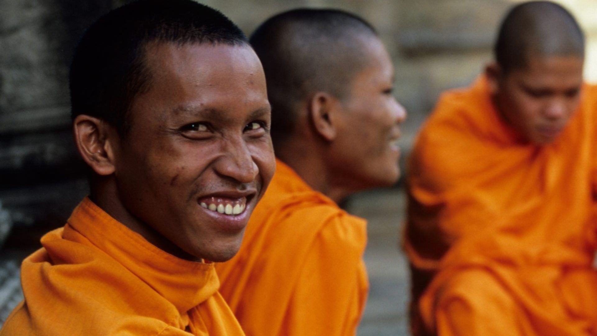 A monk in orange robes smiles to camera with two monks in the background.