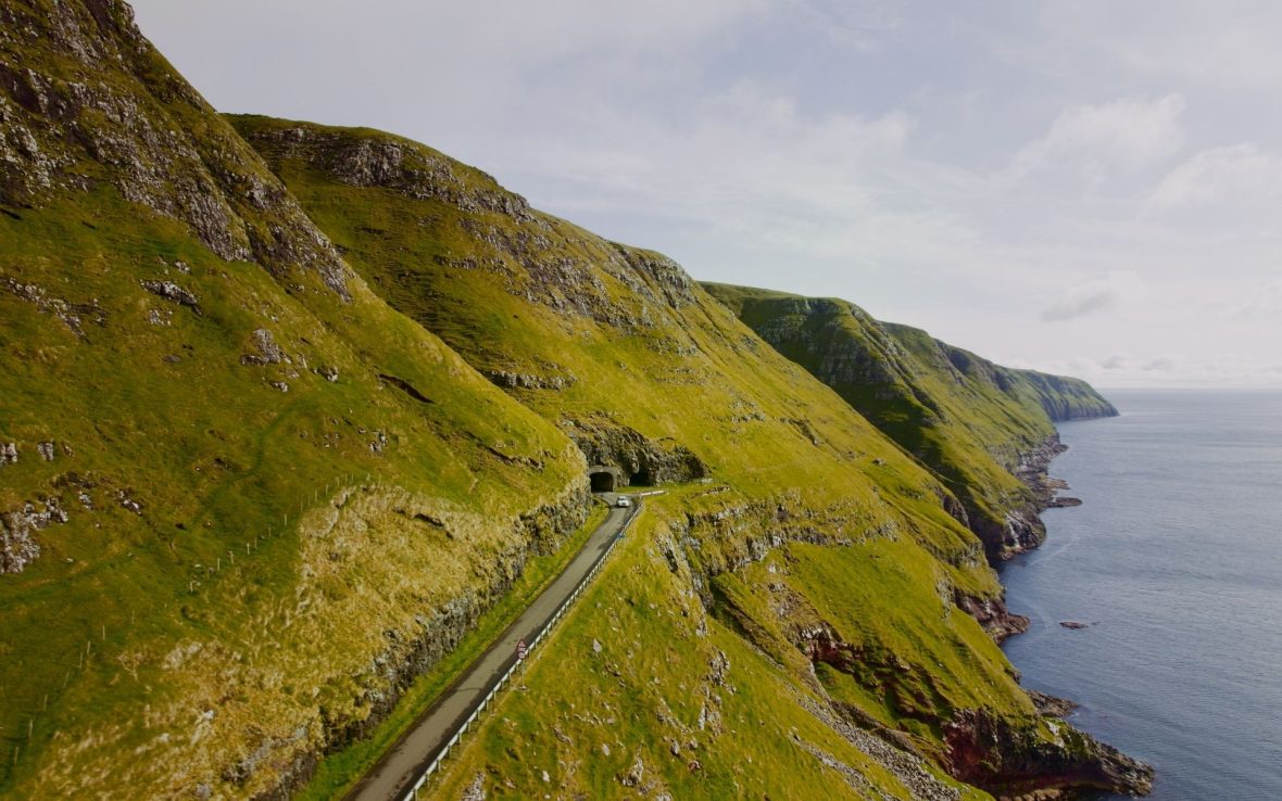 Aerial view of a car driving on a road built into the side of a grassy mountain in the Faroe Islands.