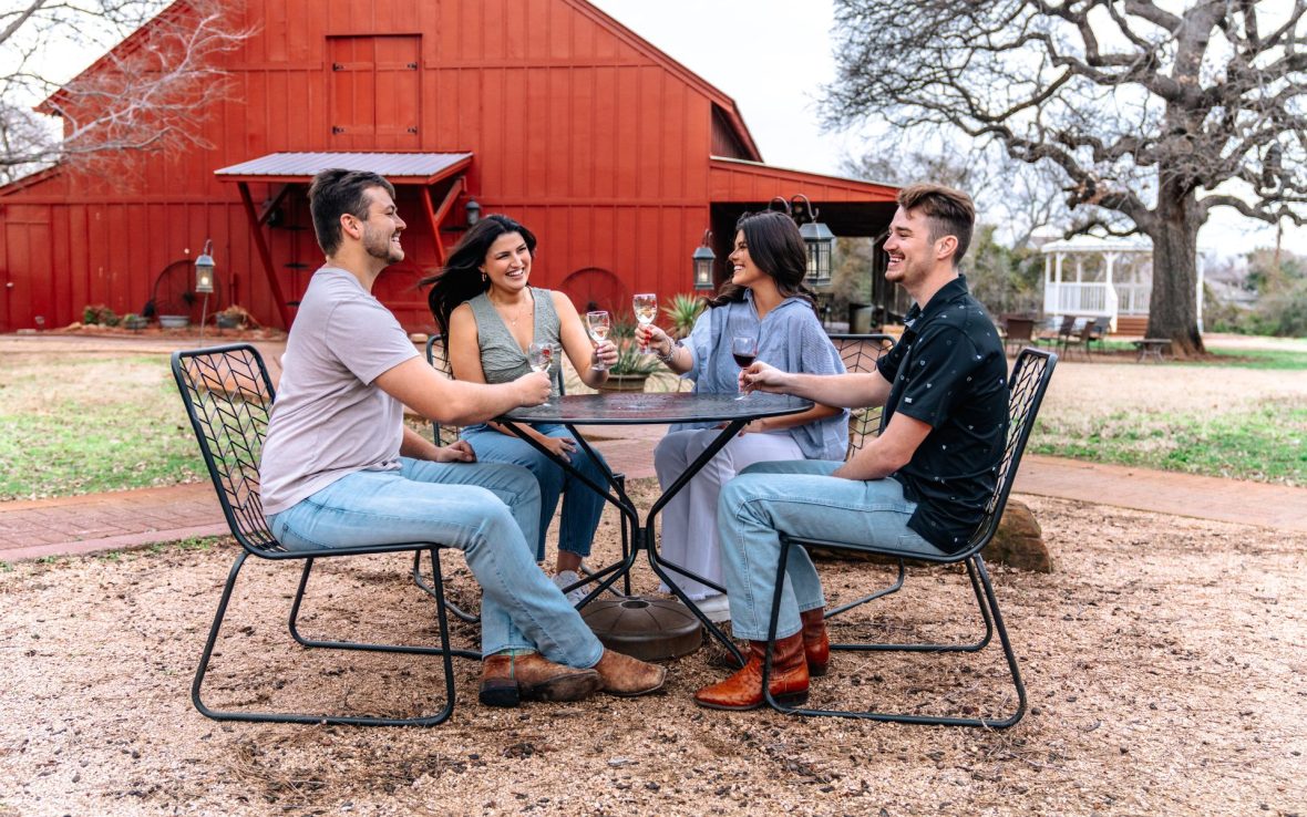 Two men and two women sit in front of a red barn drinking wine