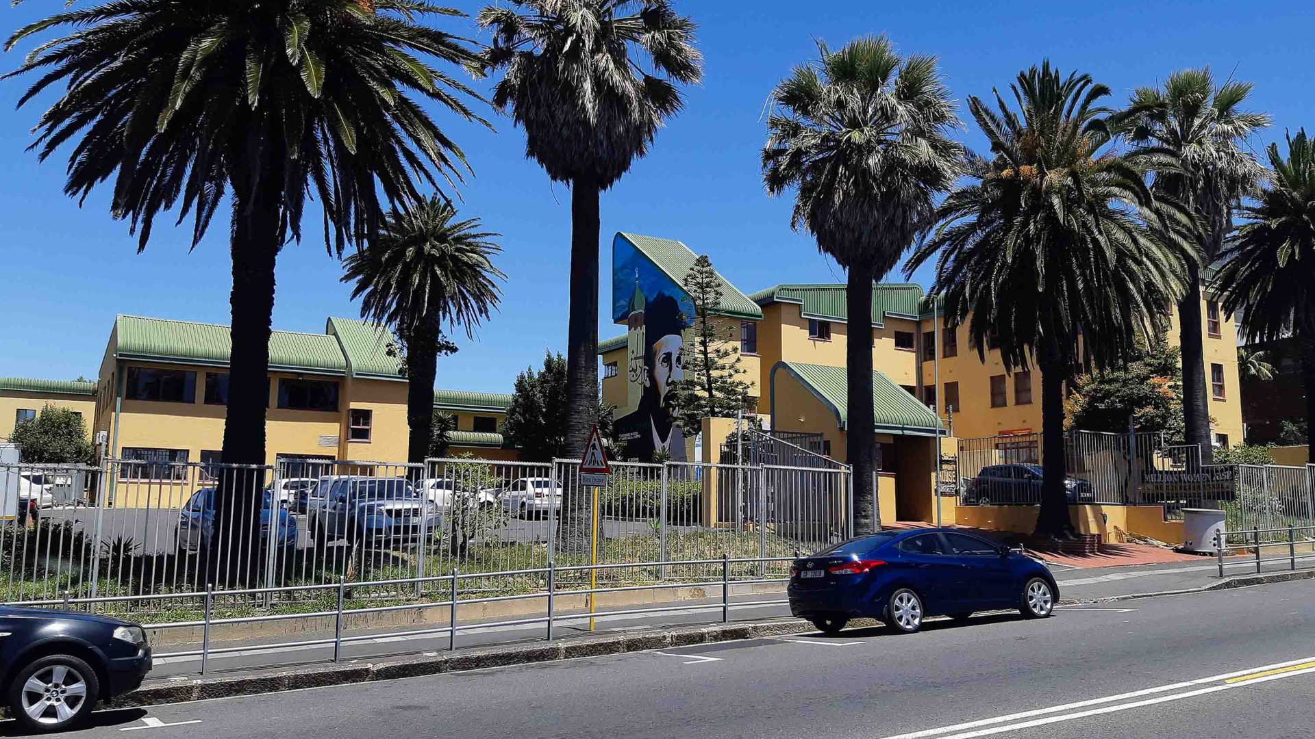 A yellow building with a mural on one wall and palm trees above.