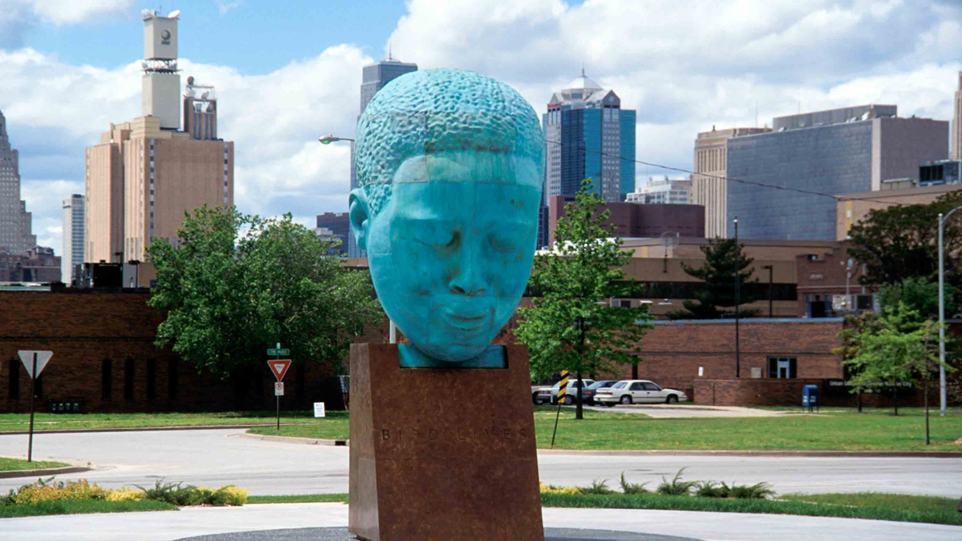 A green statue of a head stands in a square against a city backdrop