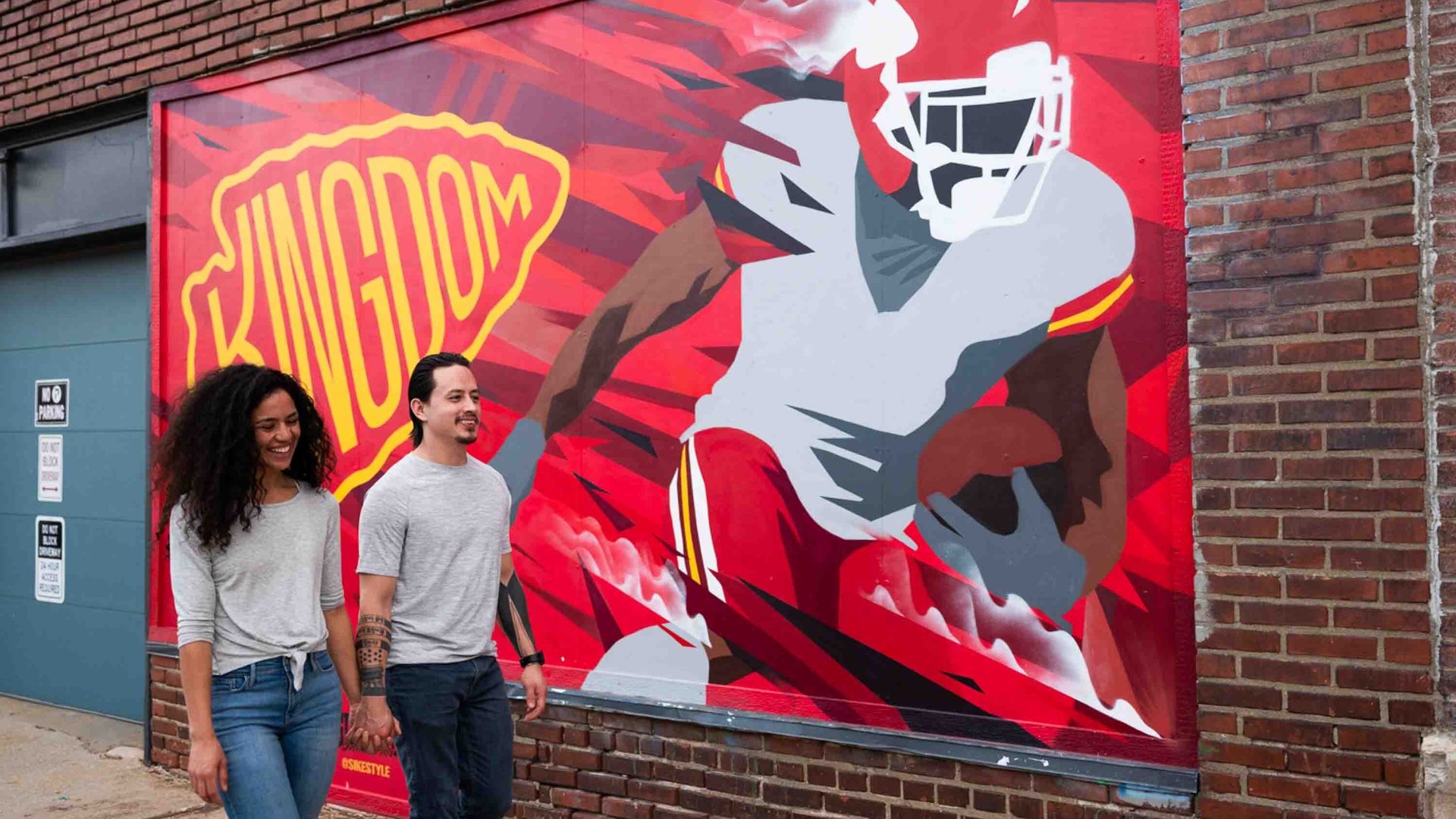 Two people walk past a striking red mural that reads 'Kingdom' alongside a graphic design of an American football player.