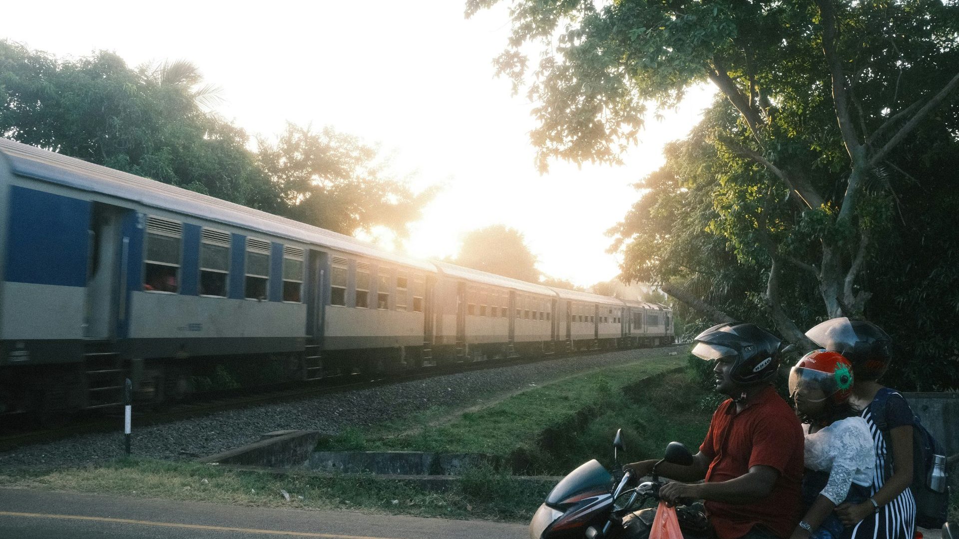 Blue and silver train passes through greenery, while a family of three ride a motorbike