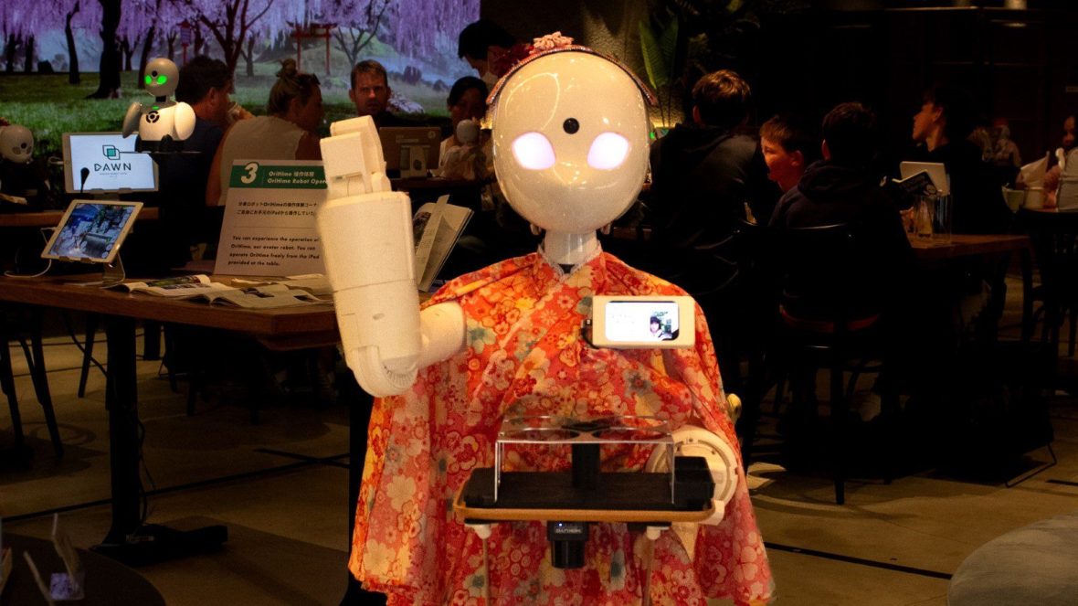 A robot avatar waves in an orange outfit waves
