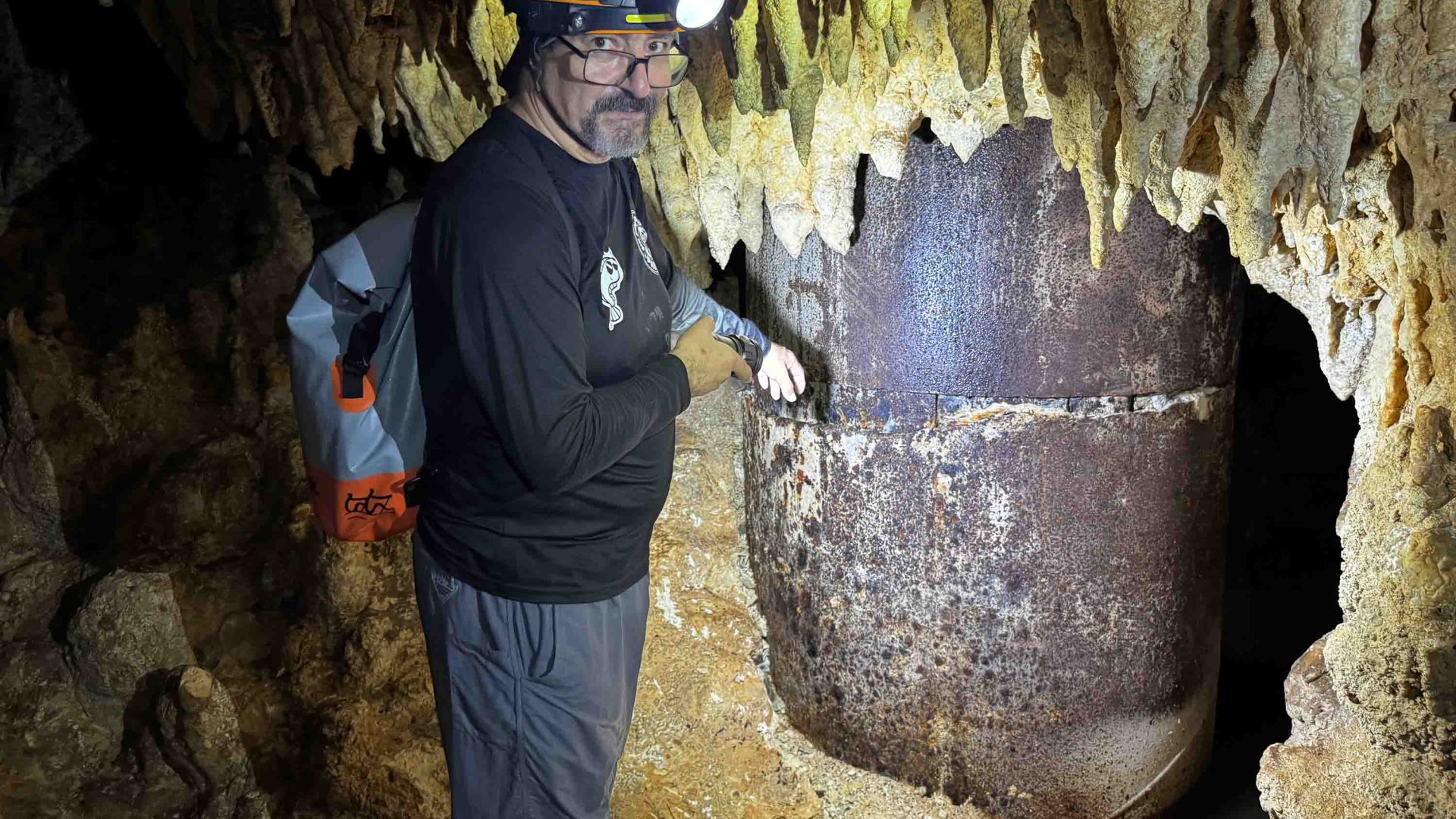 A man in a helmet with a flashlight on his head points to a train pillar in a cave.