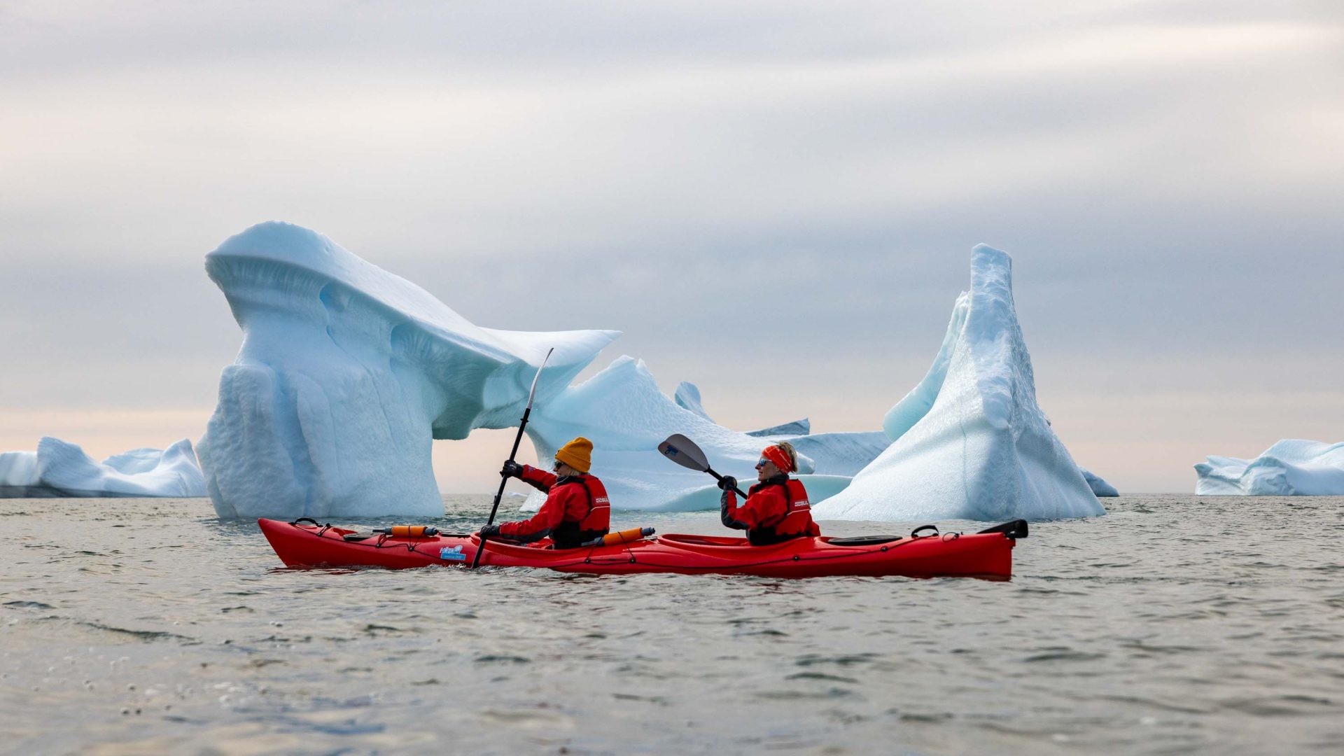 Two people in a kayak go past an iceberg.