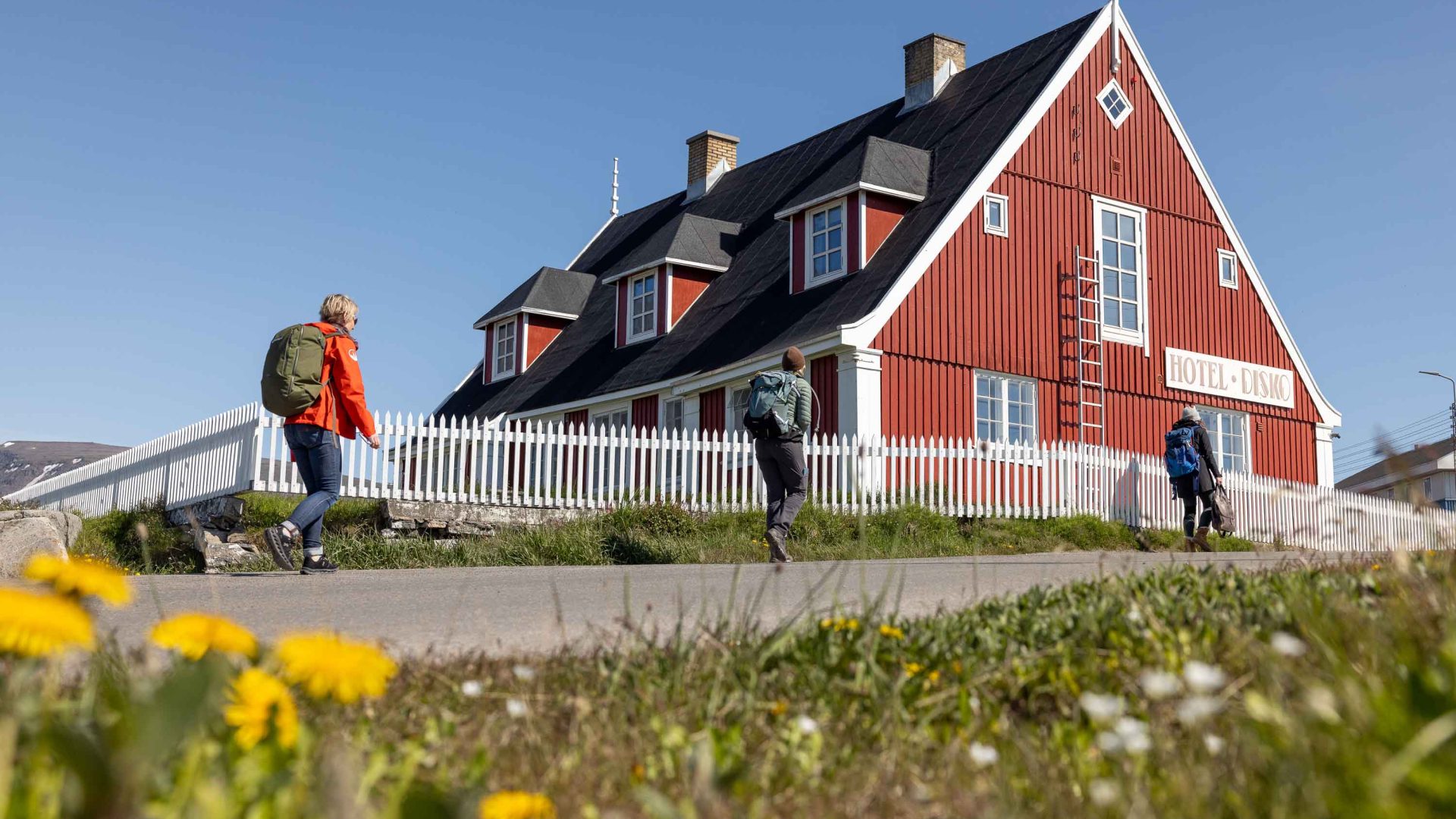 People walk past a red wooden building with yellow flowers near it.