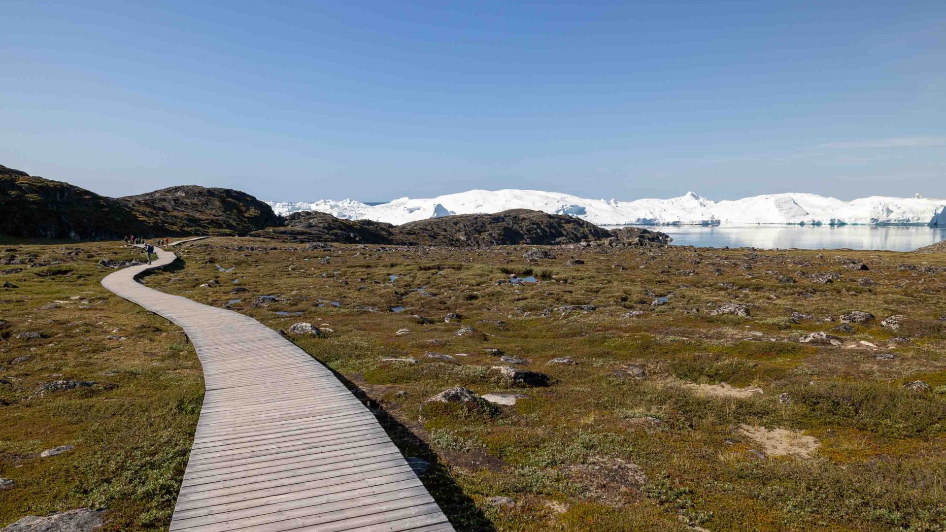A boardwalk winds up a hill toward snowy mountains.