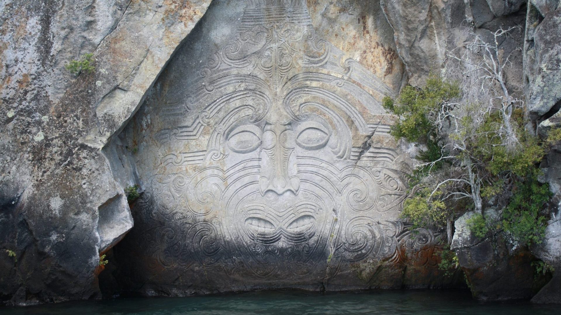 A grey cliff over water is etched with traditional Maori symbols