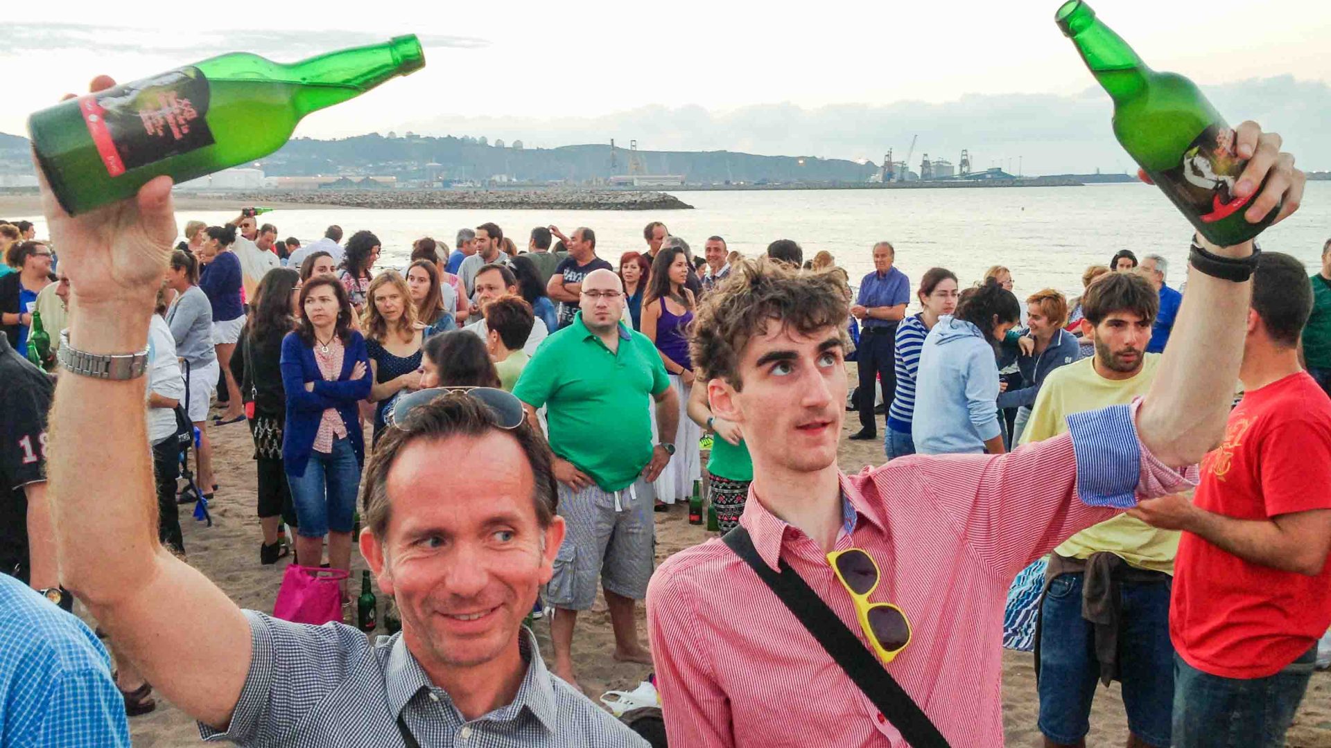 Two men pour cider surrounded by many other people at a beach.