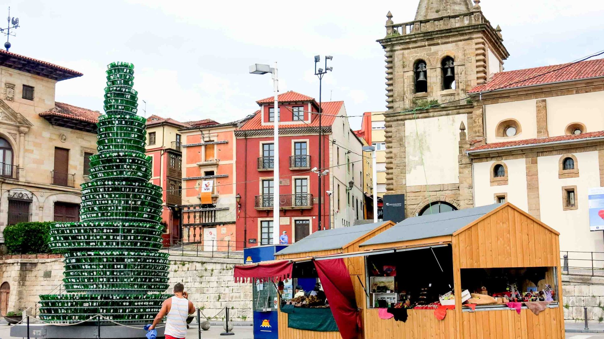 A tree made from green bottles alongside a stall and with colourful buildings nearby.