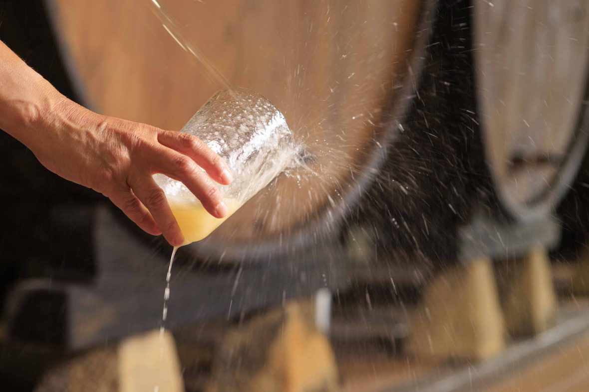 A hand holds a glass of cider that is being poured from high above.