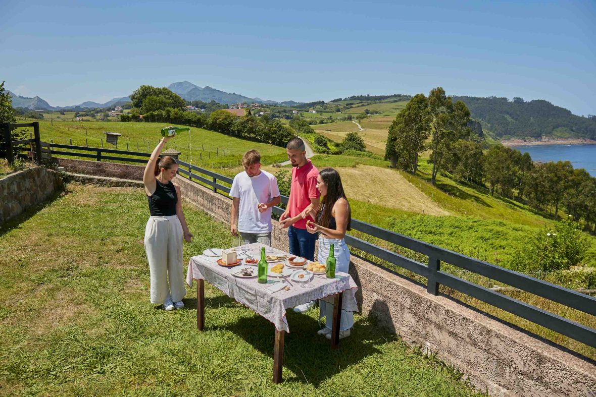 Friends stand around a table with food on it. The woman pours cider.