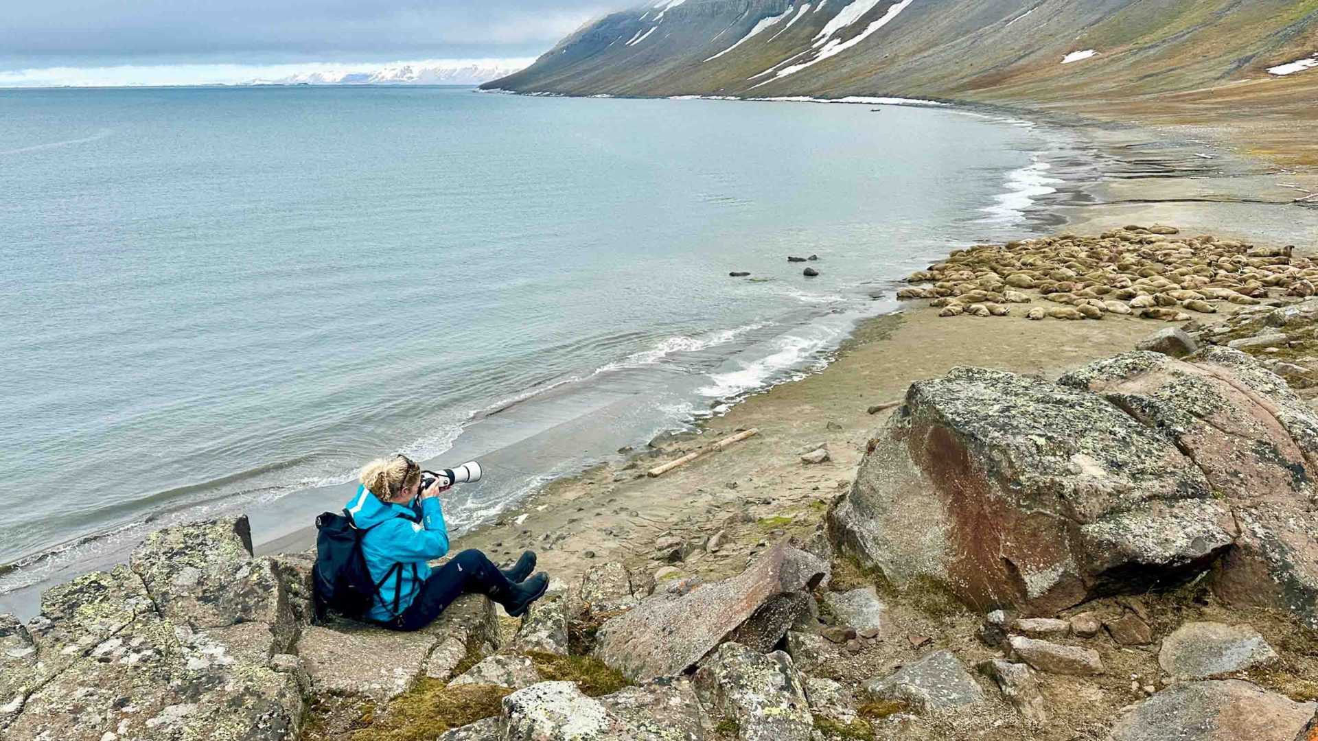 A woman sits on a rock and takes photos by the water.