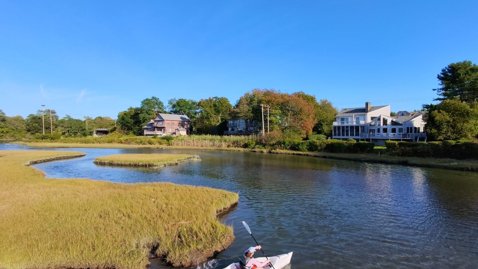 An aerial view of a woman in a light purple shirt paddles a white kayak around a grassy sandbar in the Narrow River of Narragansett, Rhode Island. Houses are visible in the distance on the other side of the river.