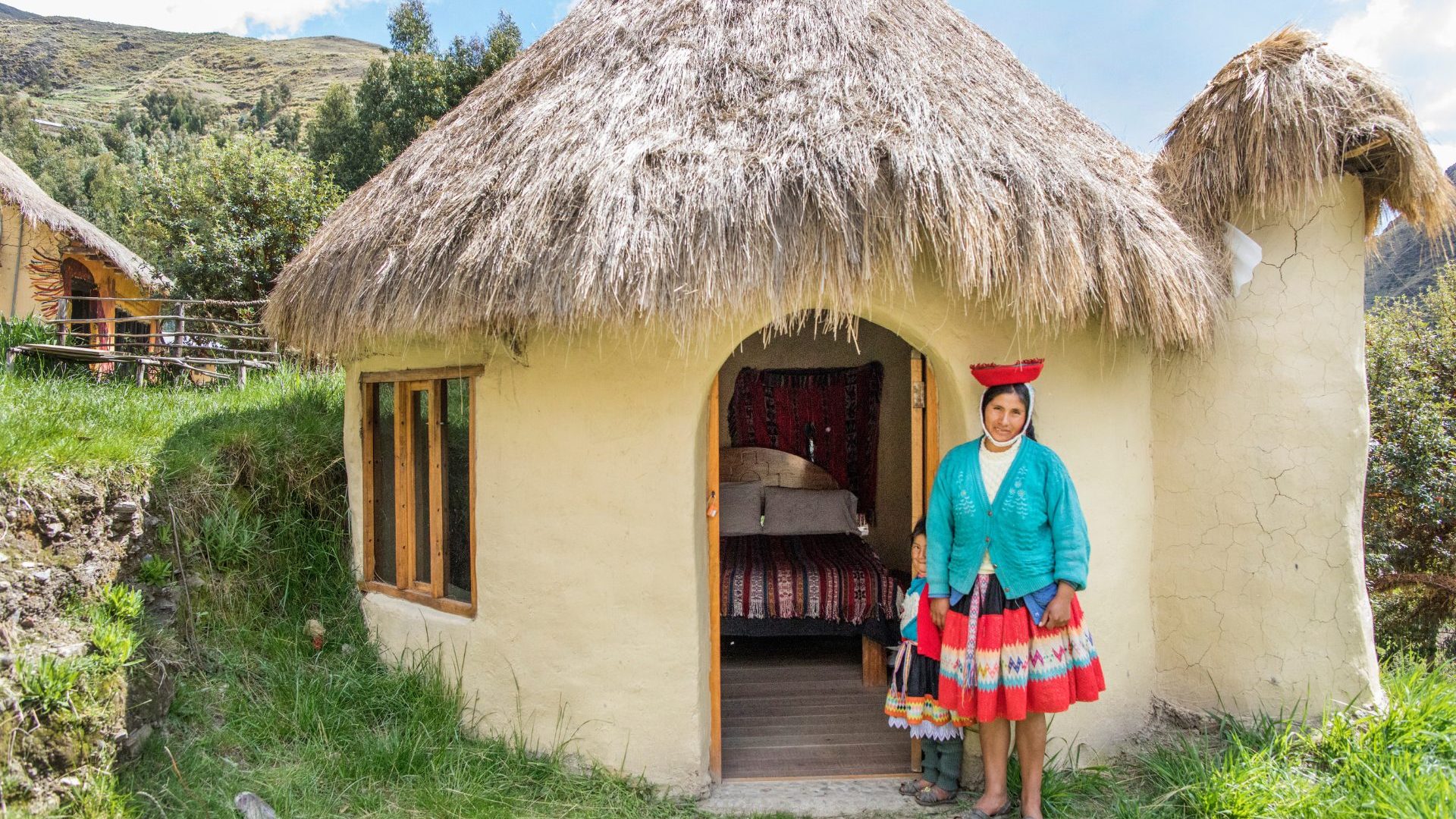 Quechua woman wearing blue and pink stands out front of a thatched roof buulding.