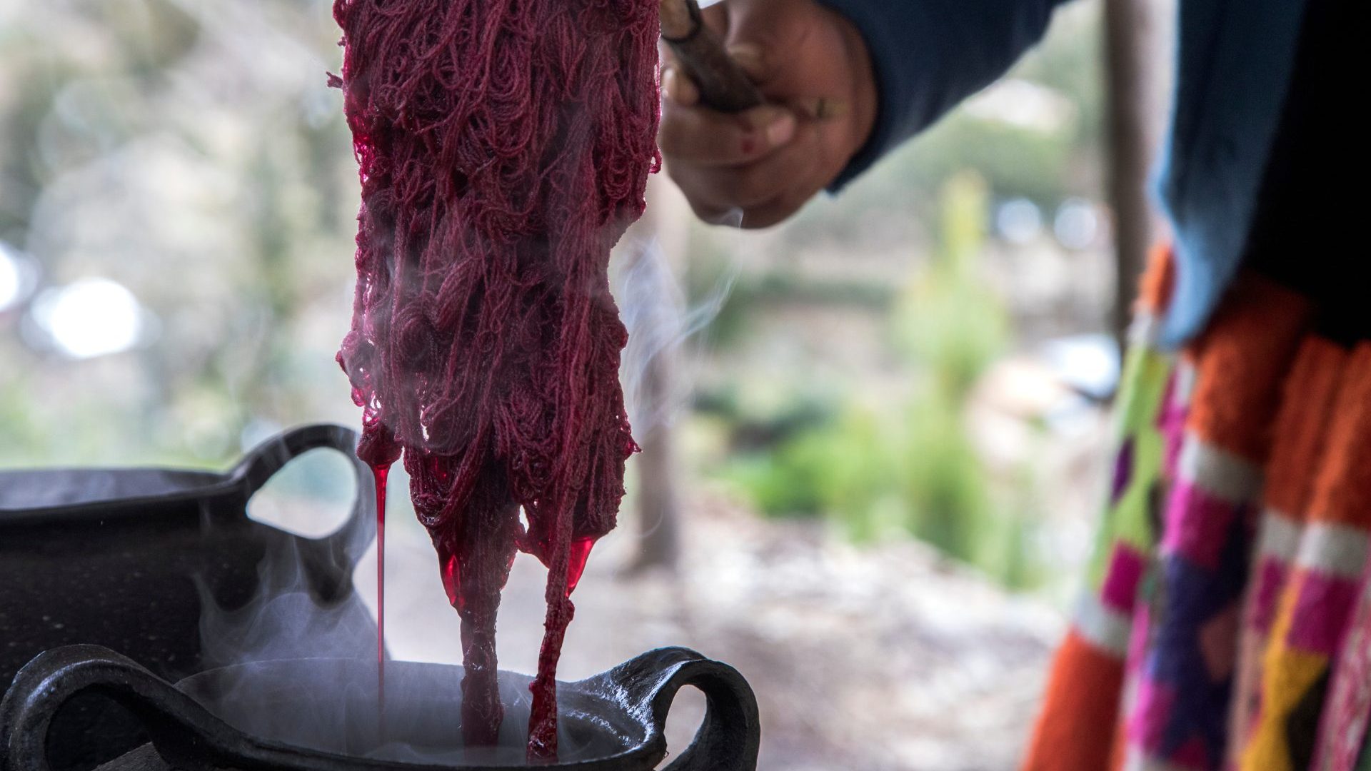 A hand pulls yarn died red out of pot.