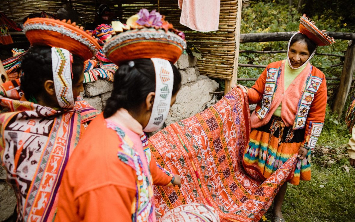 Three women wearing traditional dress hold red and pink woven fabric.