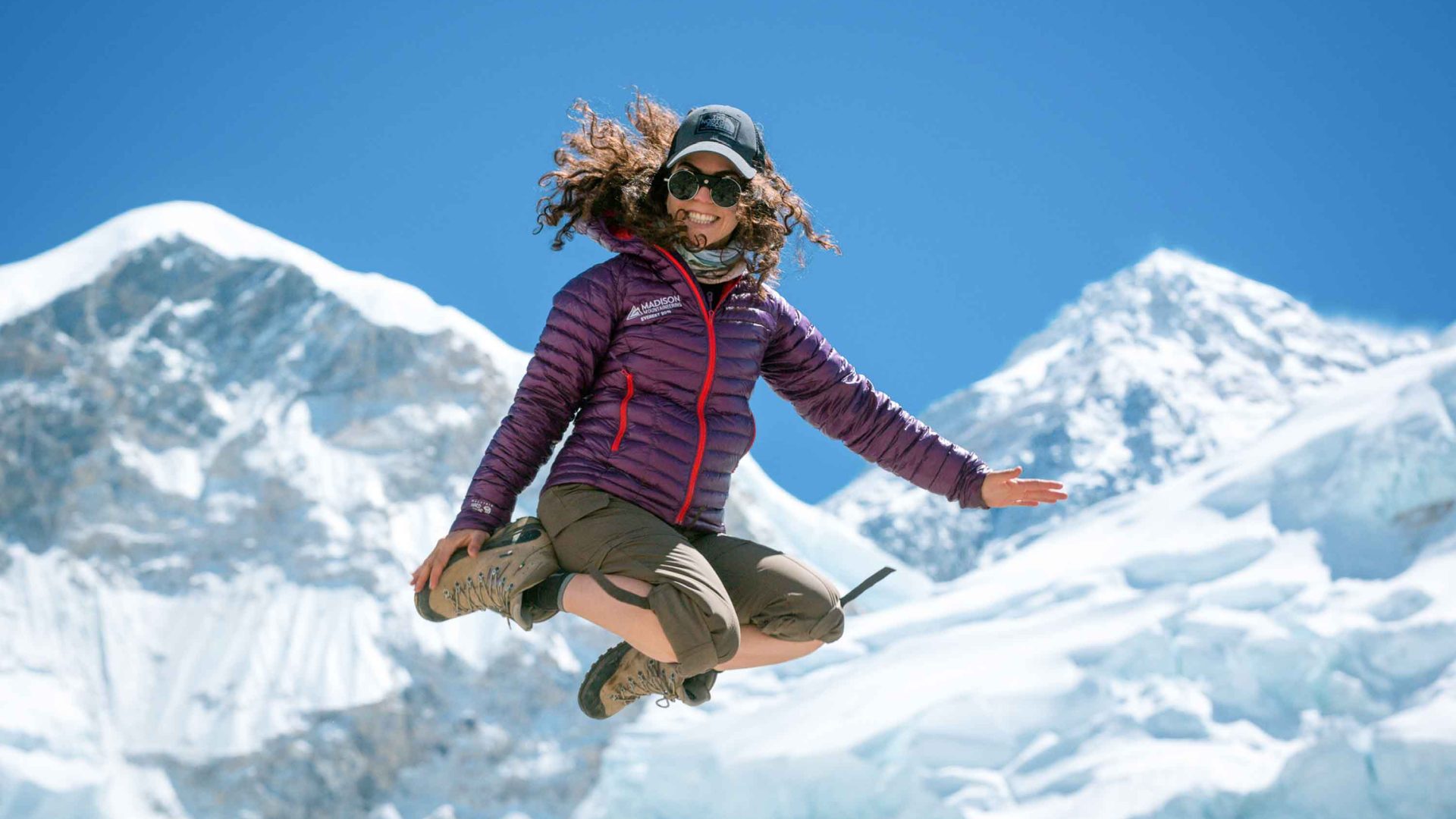 A woman in a purple down jacket jumps in the air in front of Mount Everest.