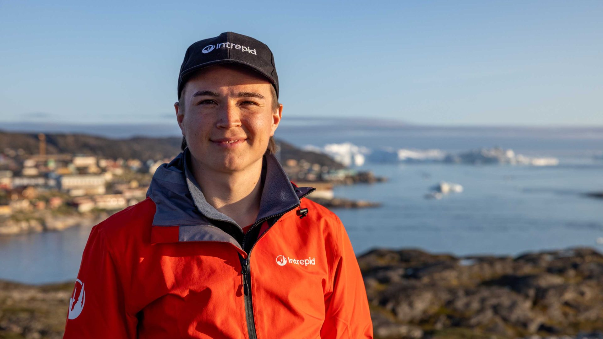 A man in red with a black har looks to camera. The sea is behind him.