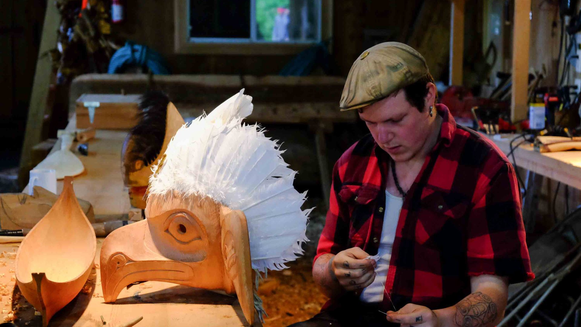A man in red and black works on a wooden mask with a white feathered head piece.
