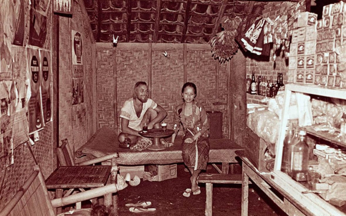 Sepia-toned photograph of male traveler sitting beside woman wearing Balinese dress at a road side stall