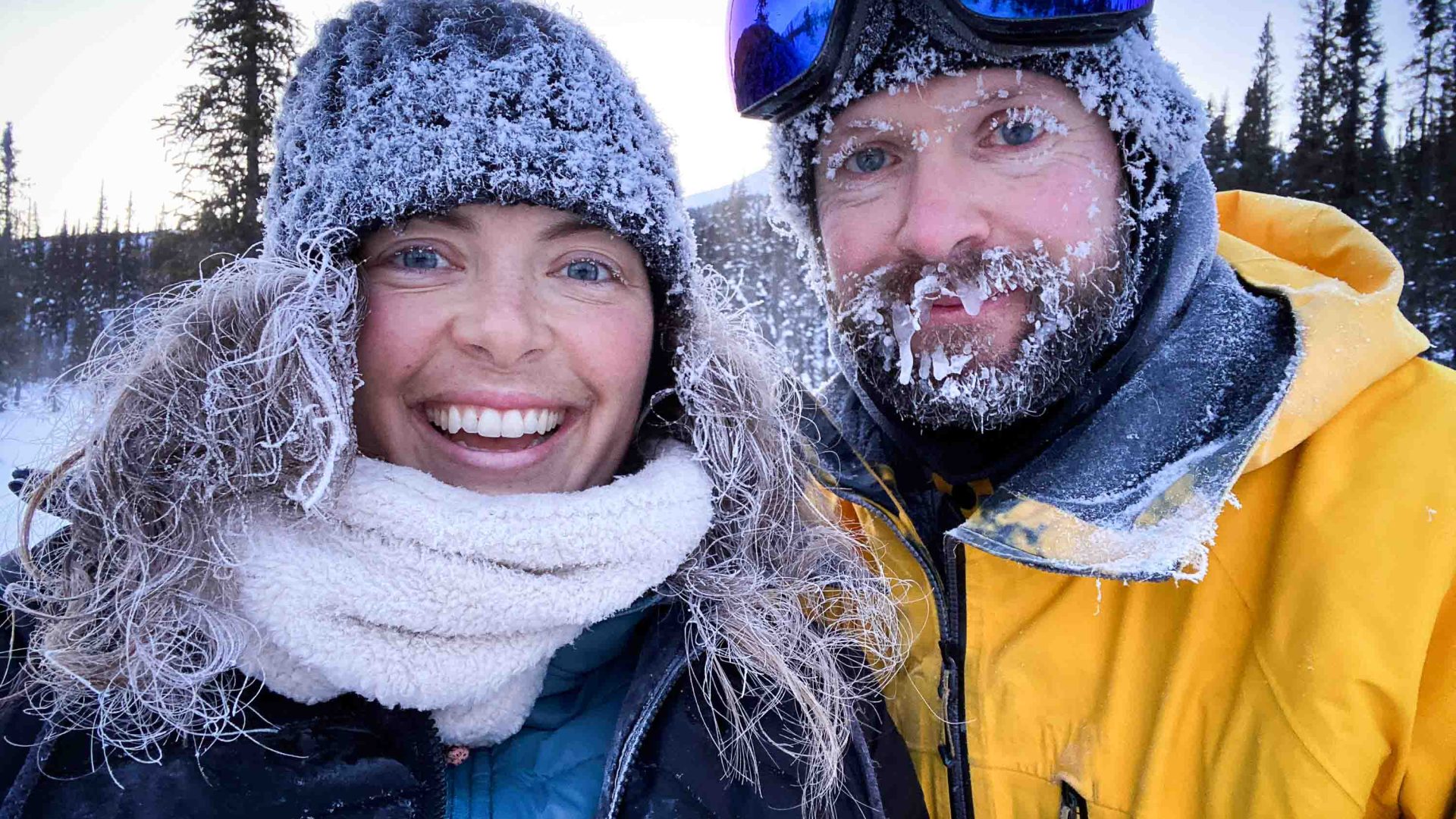 Two people smile for camera while covered in snow.