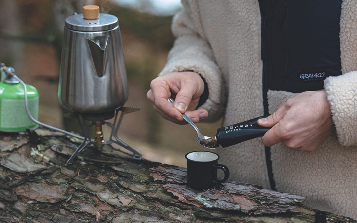 Close-up of a person's hands as they squeeze coffee paste out from a toothpaste-like tube, onto a spoon. A coffee mug sits atop a log, and to the left, a coffee pot sits atop a campstove.