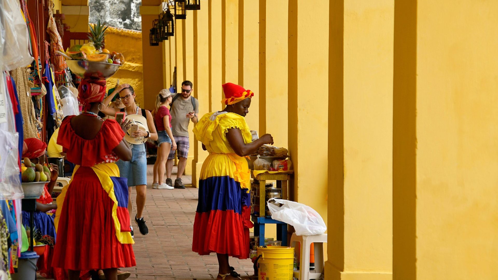 Brightly dressed women sell fruits and candies in a yellow marketplace