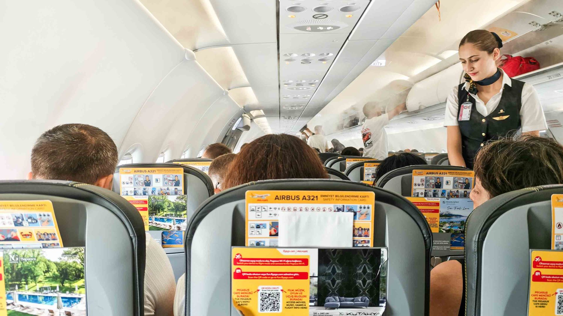 The interior of an aircraft showing safety information in the backs of seats and a female member of the cabin crew in the aisle.