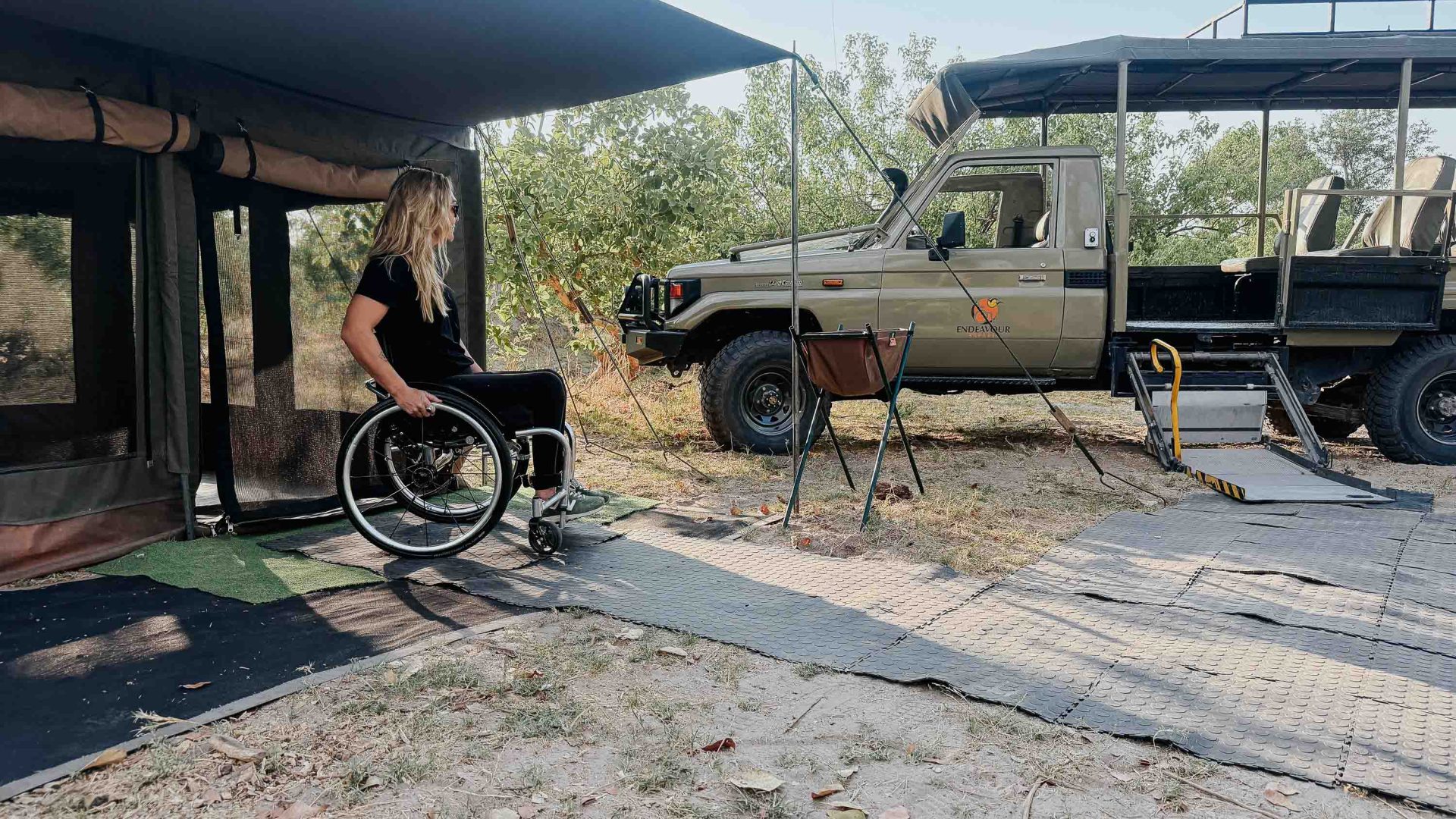 A woman wheels her chair out of her tent toward a safari vehicle.