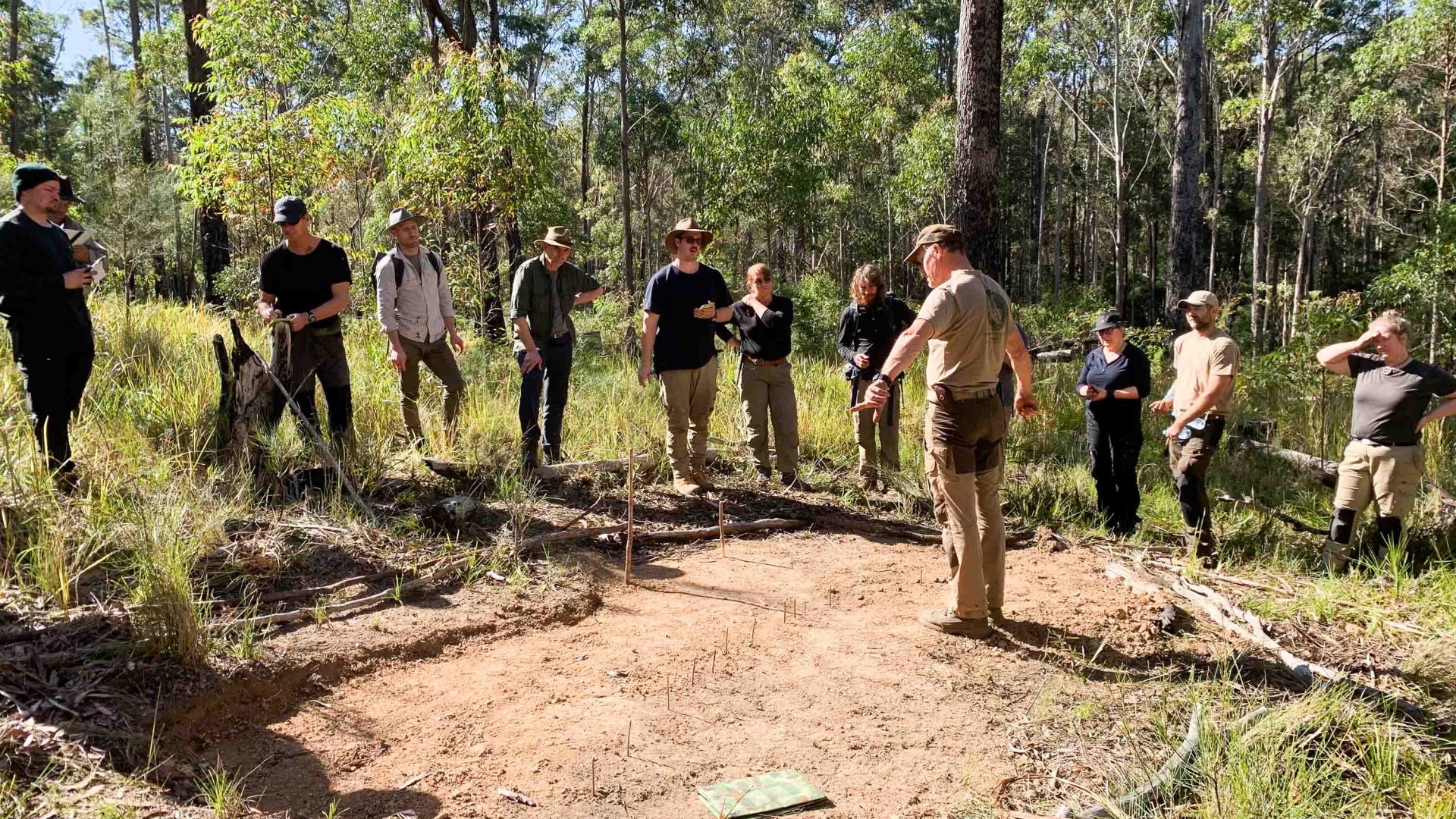 A man stands in the centre of a dirt circle and explains navigation to a group of people watching him.