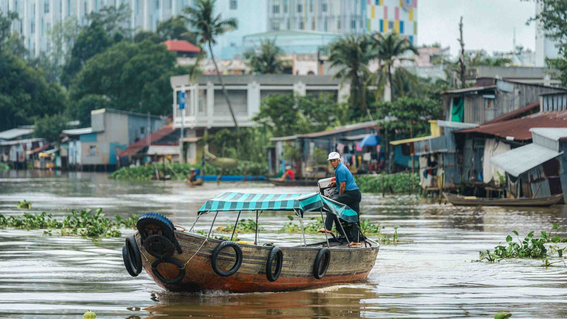 A man drives a boat along a river with houses in the background.