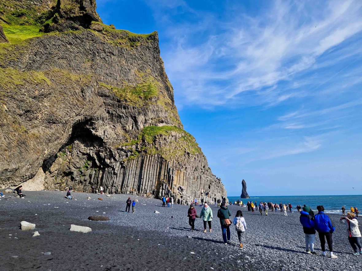 People walk along a beach with rocks jutting out of the water.