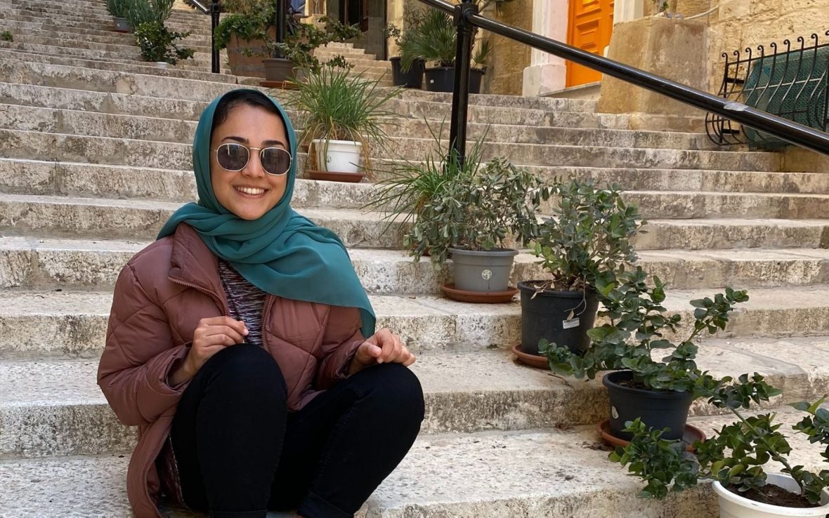 A woman wearing a hijab and sunglasses is smiling and sitting on steps.