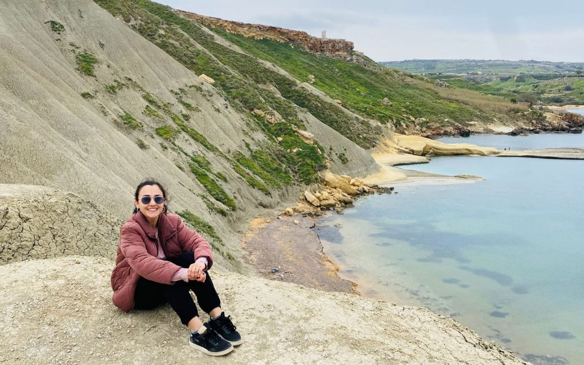 A woman sits on a rock with a rugged coastline and beach in the backdrop.