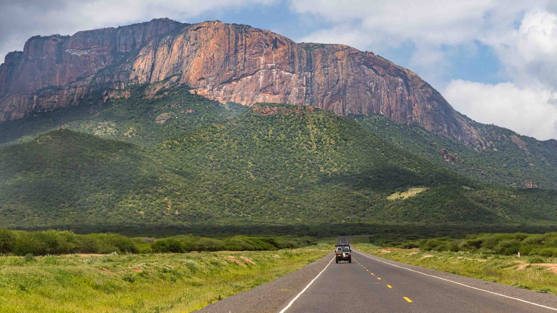 A car drives along a road away from a mountain in the distance.