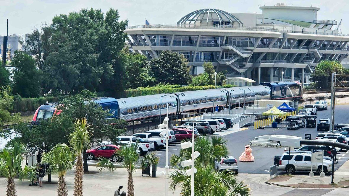 A train outside a big building with cars parked in a carpark.