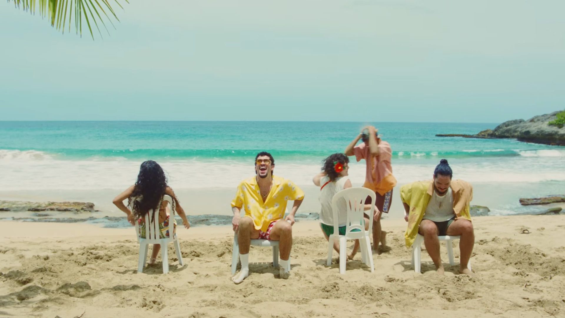 Five people play a game of musical chairs on white sand beach under blue sky.