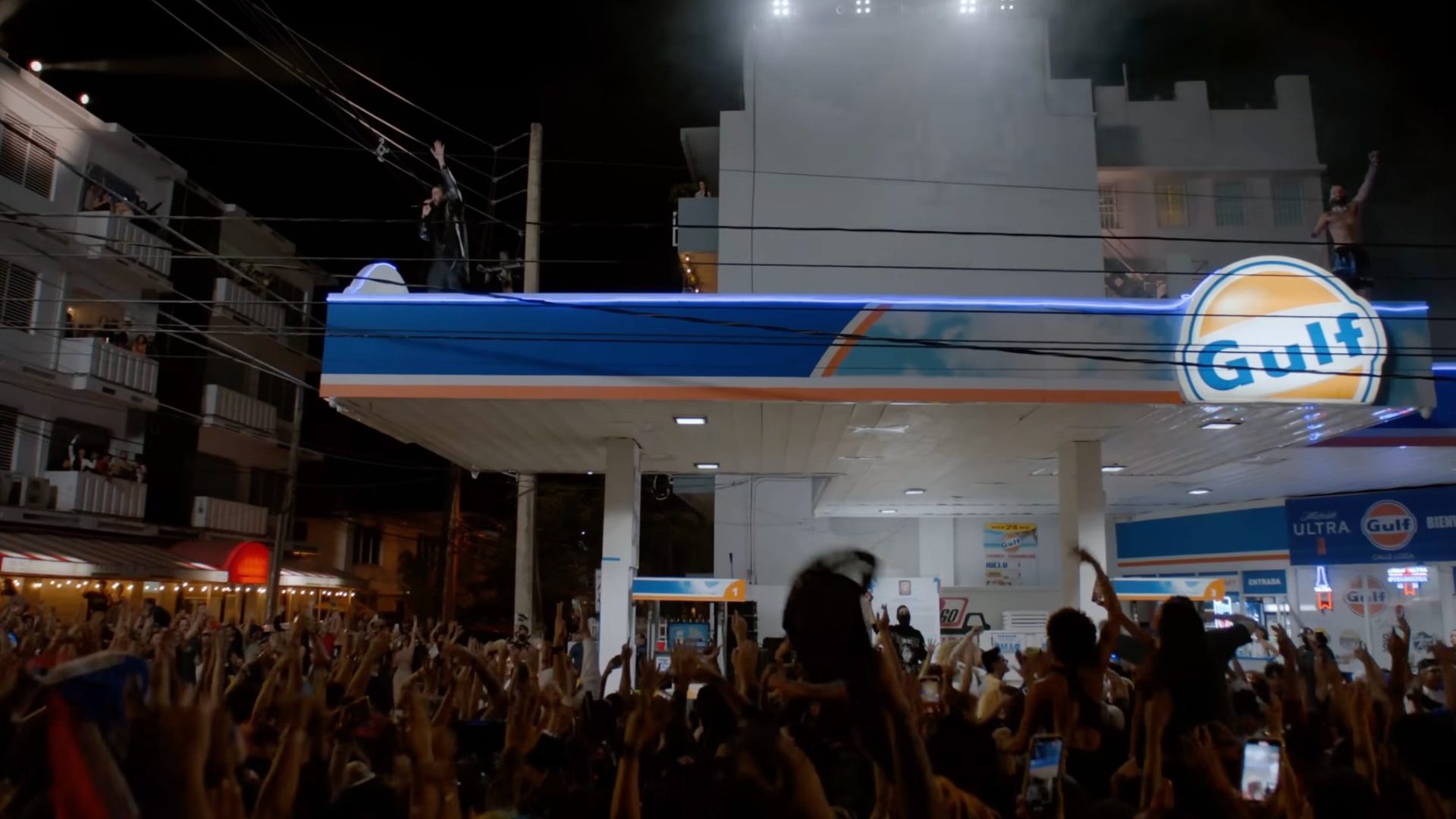 Two musicians perform on gas station roof at night to large crowd.