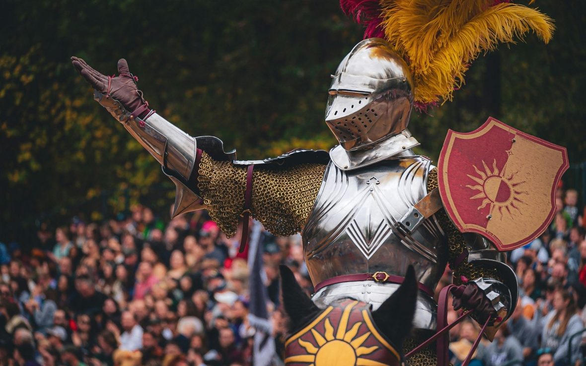 A knight in shiny armor with a feather piece on their helmet waves with a crowd in the background.