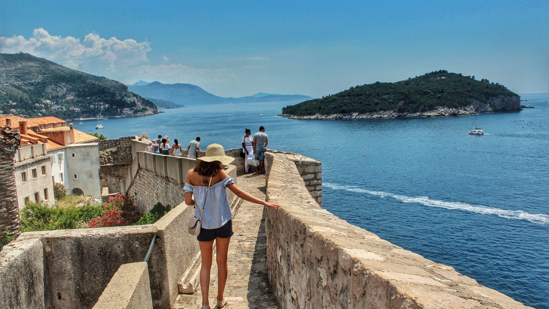 Woman walks edge of stone wall overlooking blue sea.