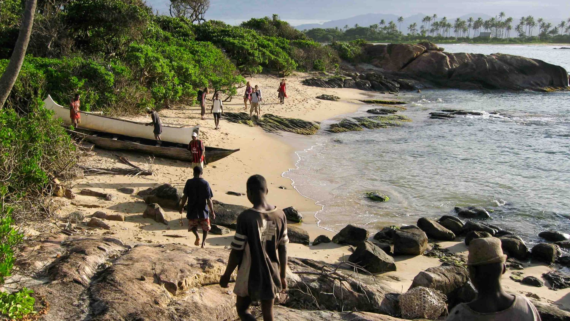 People cross a beach with rocks and wooden canoes on it.