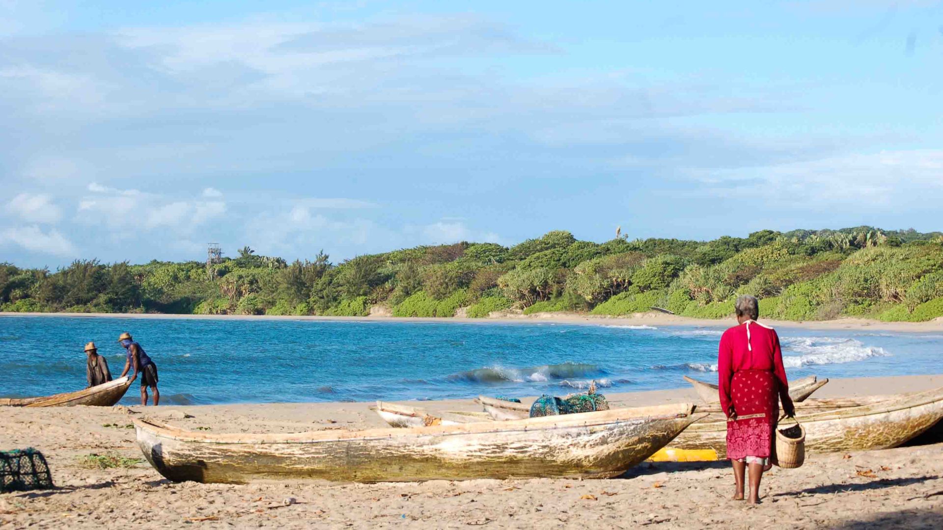 A person in red walks along a beach with wooden canoes on it.